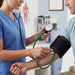 A healthcare worker in blue scrubs measures a man's blood pressure using a manual sphygmomanometer with a green tubing and arm cuff. The setting is a bright, clean clinic room.